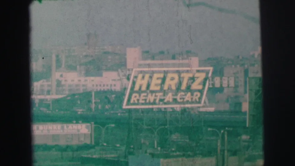 Final 1973 Lynbrook frame showing the Hertz Rent-A-Car billboard to the right within a grainy commercial streetscape.