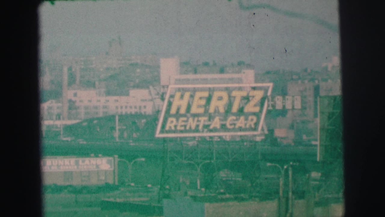Hertz Rent-A-Car billboard seen across a roadway with buildings behind it.