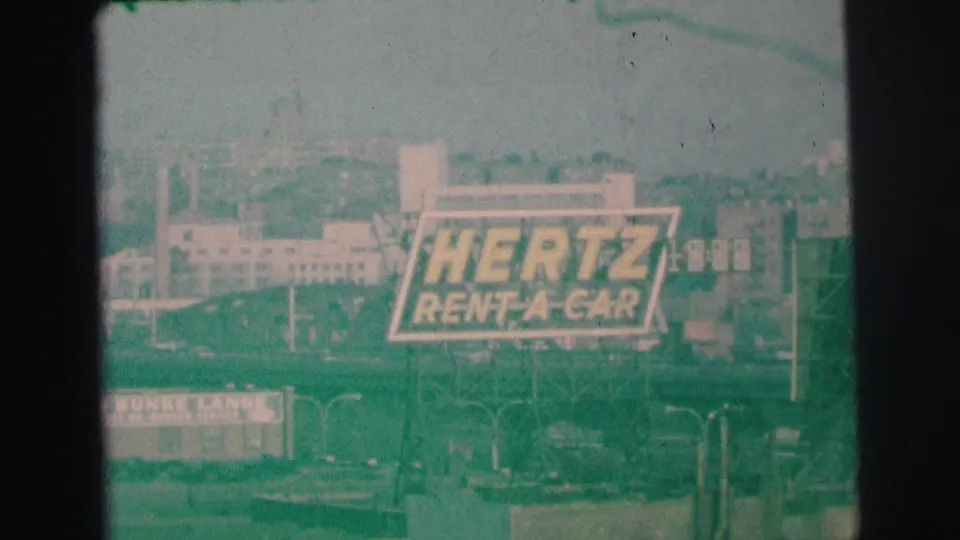 A distant Hertz Rent-A-Car billboard in Lynbrook, New York, filmed on 8mm in 1973 with hazy buildings and roadway structures in the background.