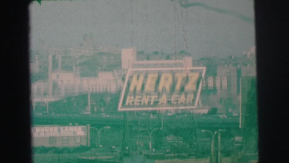 The Hertz Rent-A-Car sign remains prominent in the frame, with suburban buildings, roadway details, and faded 1973 8mm color visible around it.