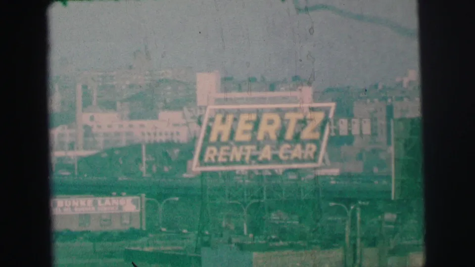 A 1973 8mm frame shows the Hertz Rent-A-Car billboard over a roadside scene in Lynbrook, with a partially visible lower sign, vehicles, poles, and aged-film texture.