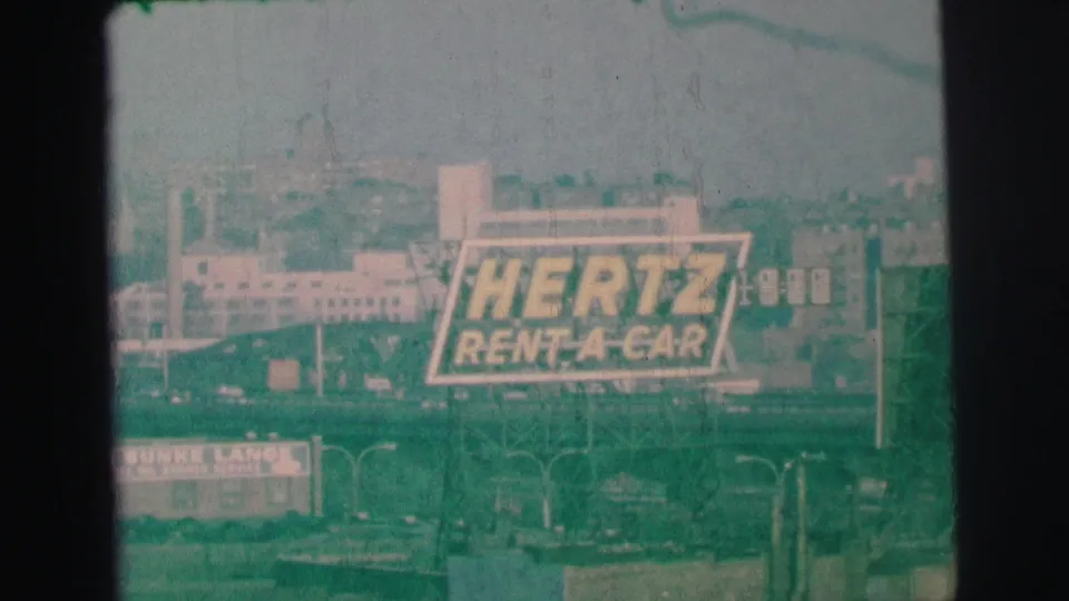 The final frame holds on the Hertz Rent-A-Car billboard in Lynbrook, New York, with hazy background buildings, a roadside foreground, and the soft faded look of 1973 8mm film.