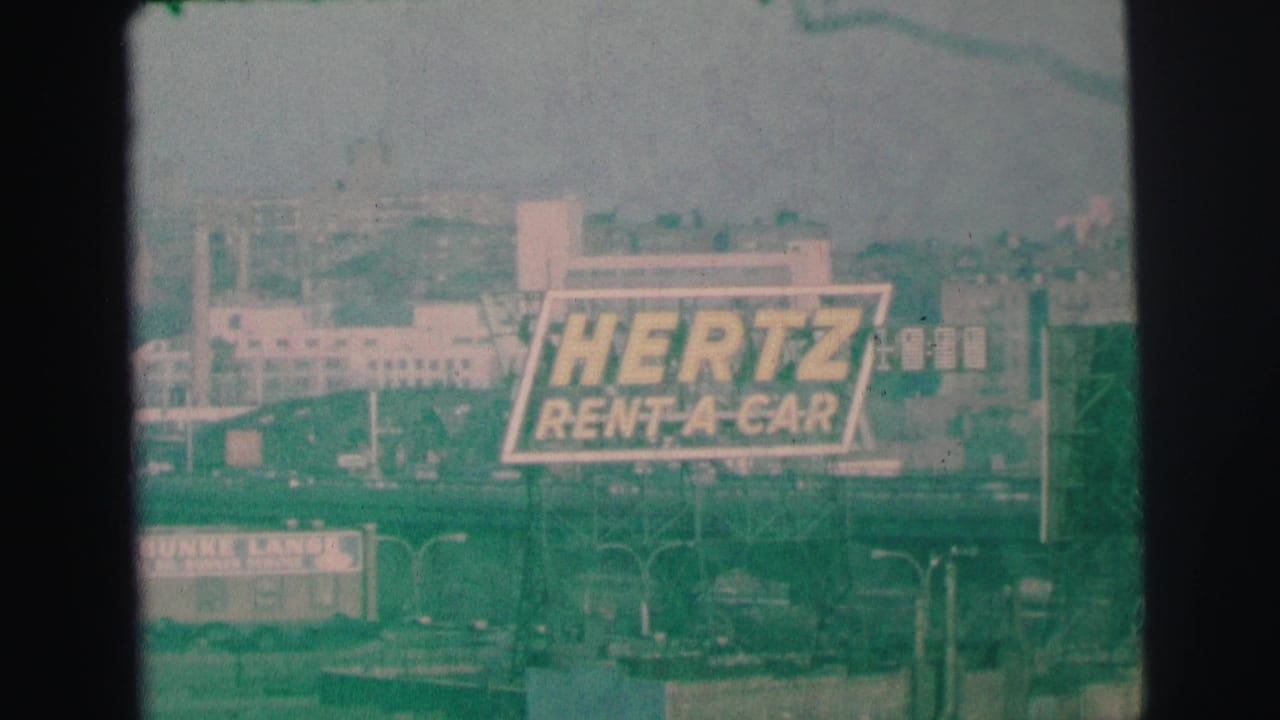 Large Hertz Rent-A-Car billboard above a distant low-rise streetscape.