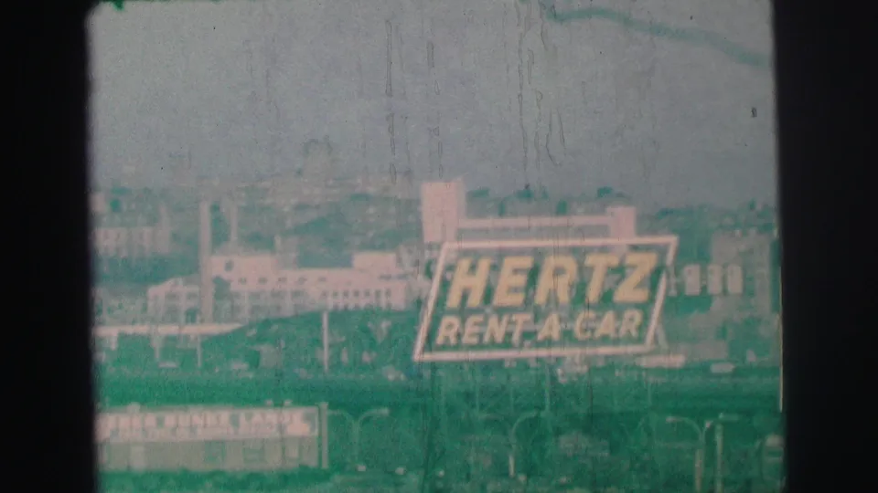A green-tinted 1973 frame shows the Hertz Rent-A-Car sign above buildings, with additional street signage near the lower edge.