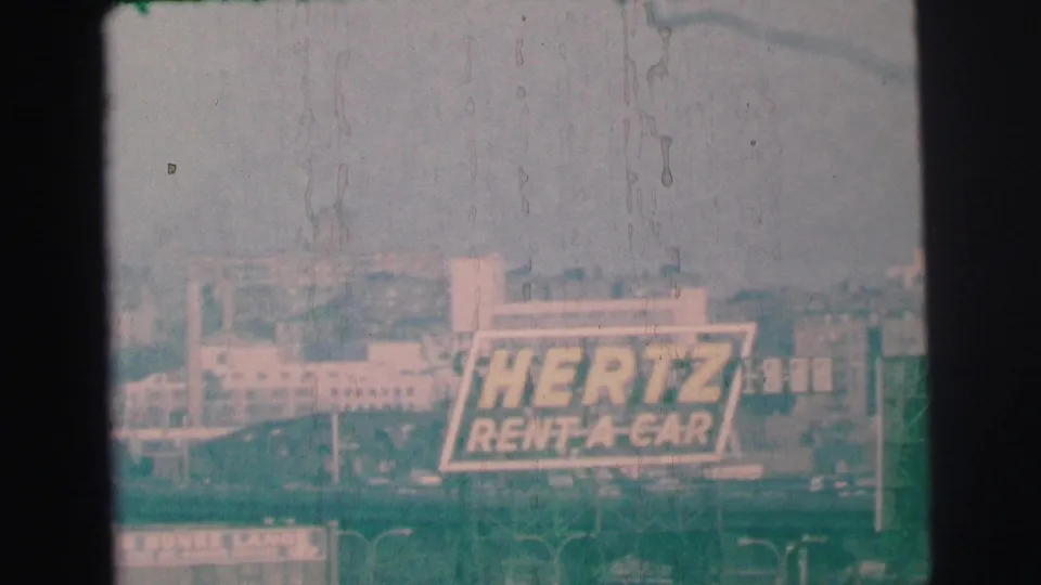 The final 8mm frame keeps the Hertz Rent-A-Car billboard in view over a soft, green-shifted Lynbrook streetscape.