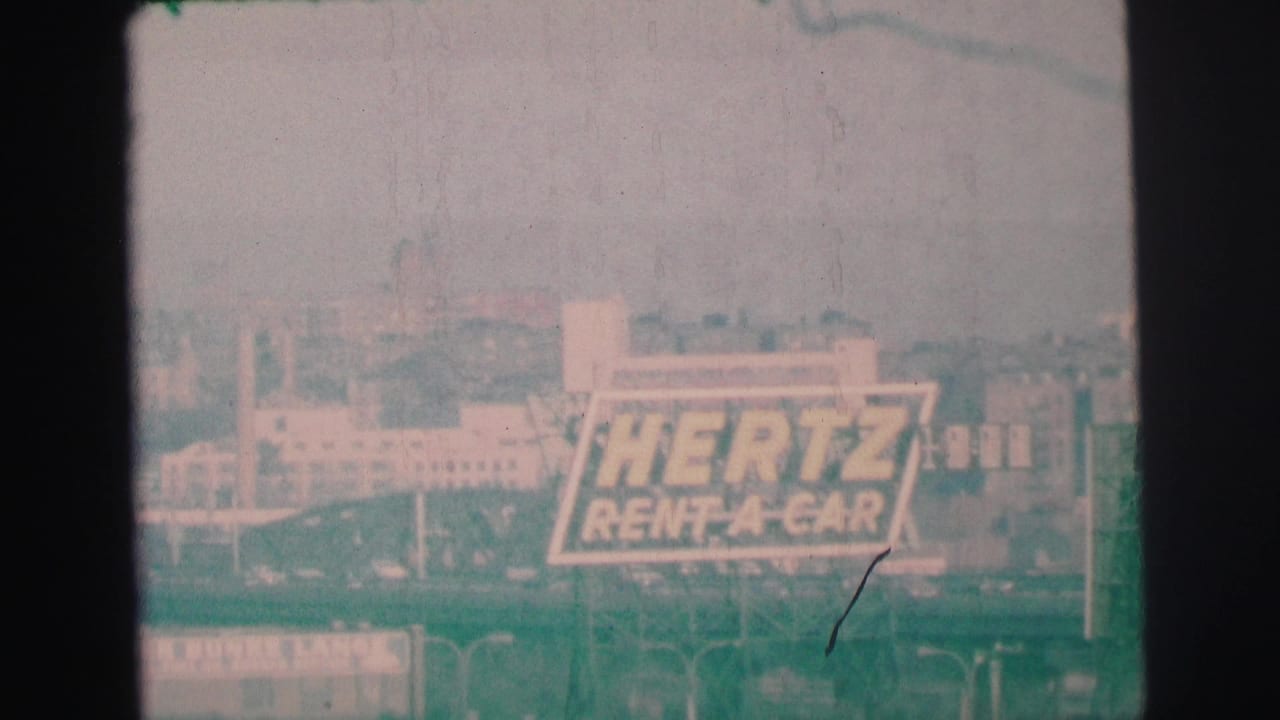Hazy cityscape with a large Hertz Rent-A-Car sign and dark 8mm frame borders.