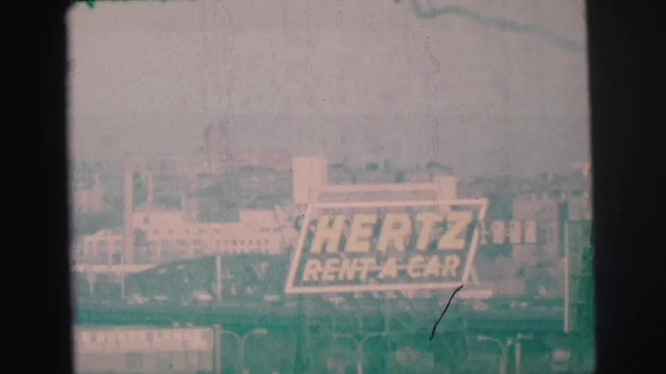 1973 8mm view in Lynbrook, New York, showing a large Hertz Rent-A-Car sign against hazy buildings and distant stacks.