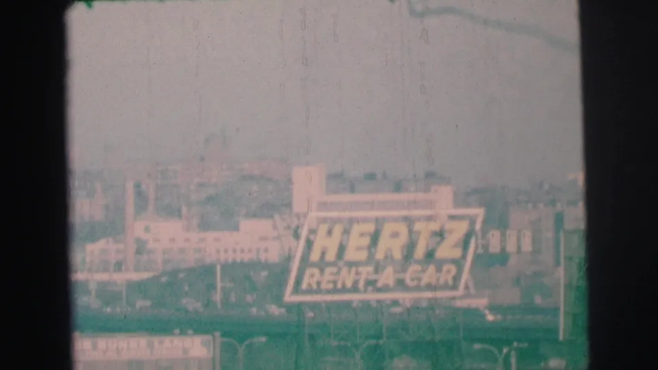 Lynbrook, New York home-movie frame with the Hertz Rent-A-Car sign centered amid low buildings and soft 8mm haze.