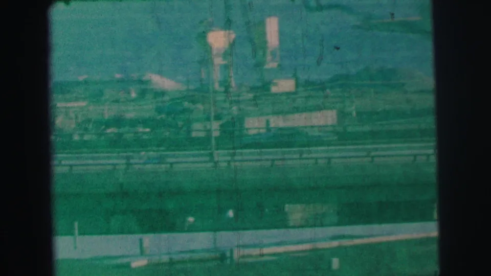 Final 8mm frame from the Lynbrook roadside view, with the Hertz sign less distinct behind a pole, barriers, and green film tint.