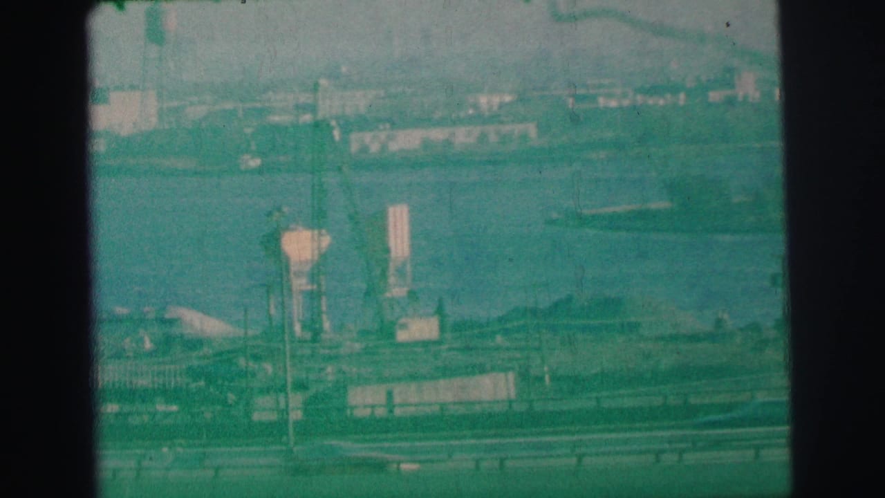 1973 Lynbrook, New York 8mm frame showing freeway traffic, guardrails, bay water, and distant industrial shoreline structures.