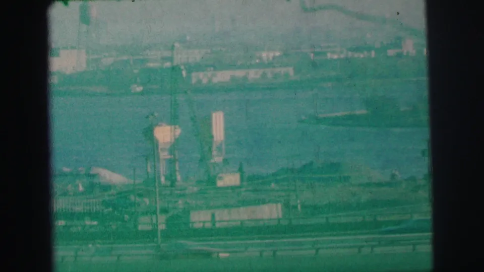 1973 Lynbrook, New York 8mm frame showing freeway traffic, guardrails, bay water, and distant industrial shoreline structures.