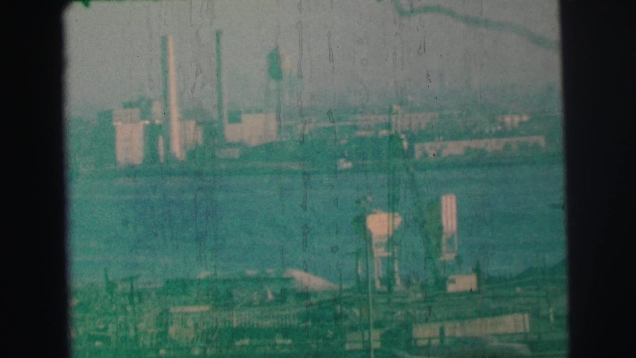 1973 Lynbrook, New York 8mm frame showing a distant industrial shoreline with smokestacks, a water tower, and dock structures through haze.