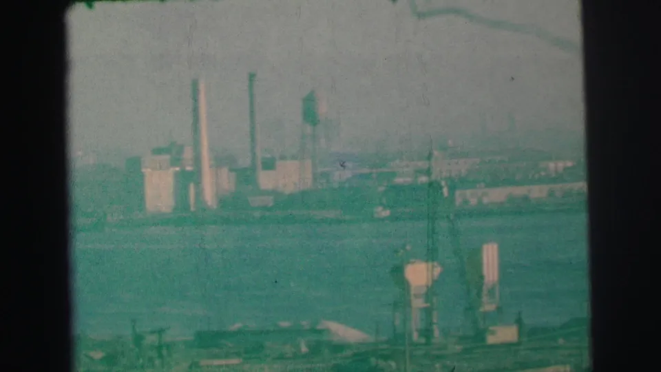 1973 Lynbrook-area waterfront view with factory smokestacks, a water tower, cranes, and low industrial buildings across the water.