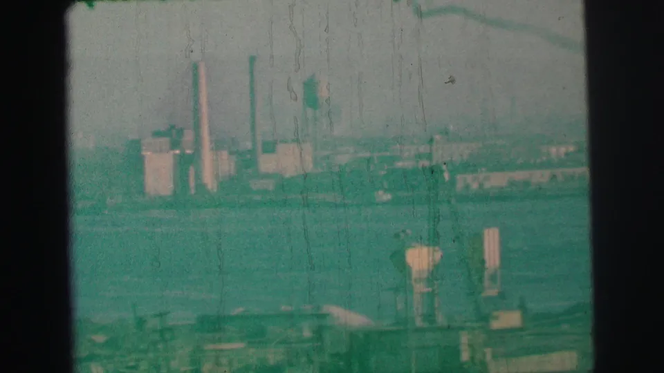 Hazy 1973 Lynbrook waterfront frame with distant smokestacks, a water tower, foreground dock equipment, and visible film wear.