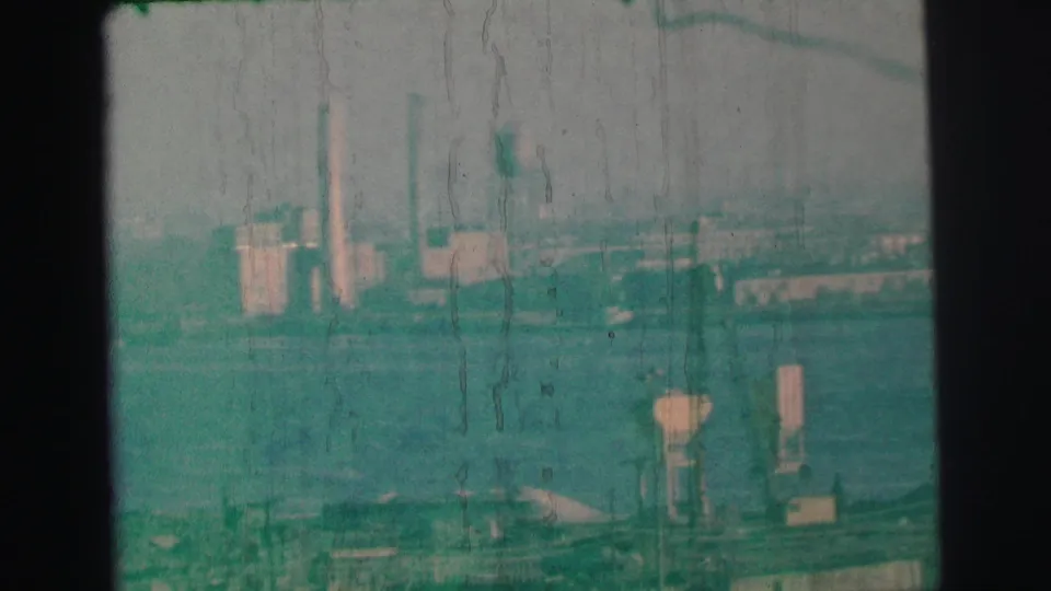 1973 Lynbrook, New York 8mm frame showing a distant industrial waterfront with smokestacks, cranes, and a water tower across the water.