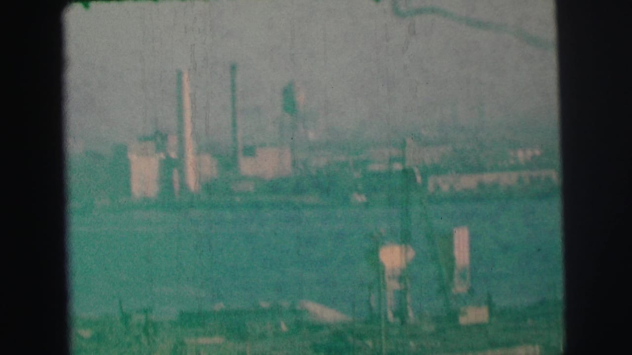 1973 Lynbrook-area 8mm frame showing a distant industrial waterfront with smokestacks, water towers, haze, and green film tint.