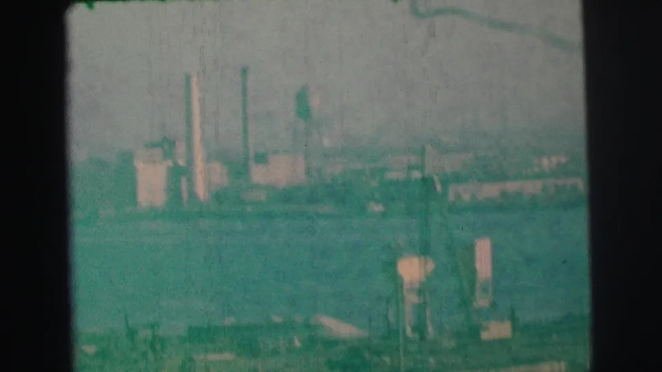 1973 Lynbrook-area 8mm frame showing a distant industrial waterfront with smokestacks, water towers, haze, and green film tint.