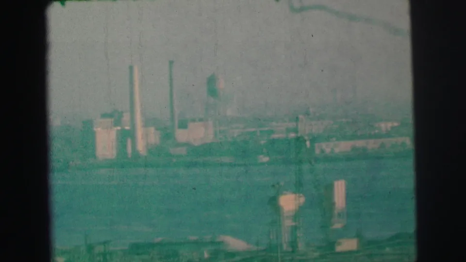 Distant Lynbrook, New York industrial shoreline in 1973, with tall stacks and a water tower seen through soft 8mm haze.