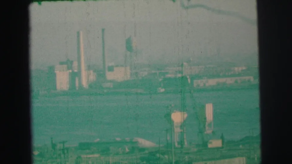 Final 1973 Lynbrook 8mm frame holding on a distant industrial riverbank with smokestacks, water towers, haze, and film grain.