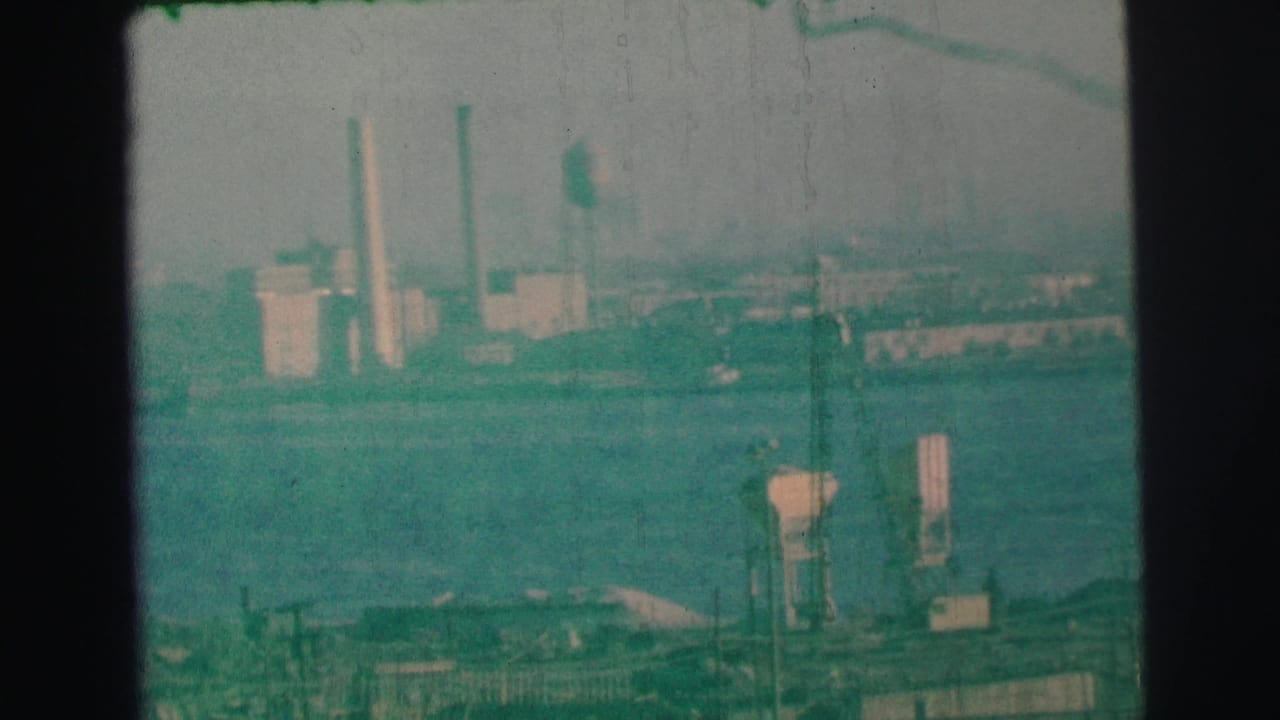 1973 Lynbrook-area home-movie view of a hazy industrial shoreline across water, with smokestacks and foreground structures.