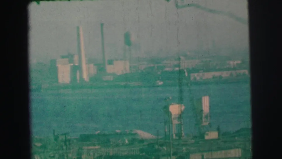 1973 Lynbrook-area home-movie view of a hazy industrial shoreline across water, with smokestacks and foreground structures.
