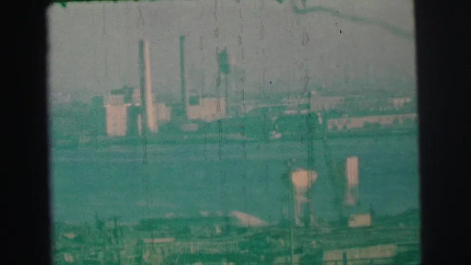 Hazy 1973 8mm view near Lynbrook, New York, showing industrial buildings and tall stacks beyond a waterfront.