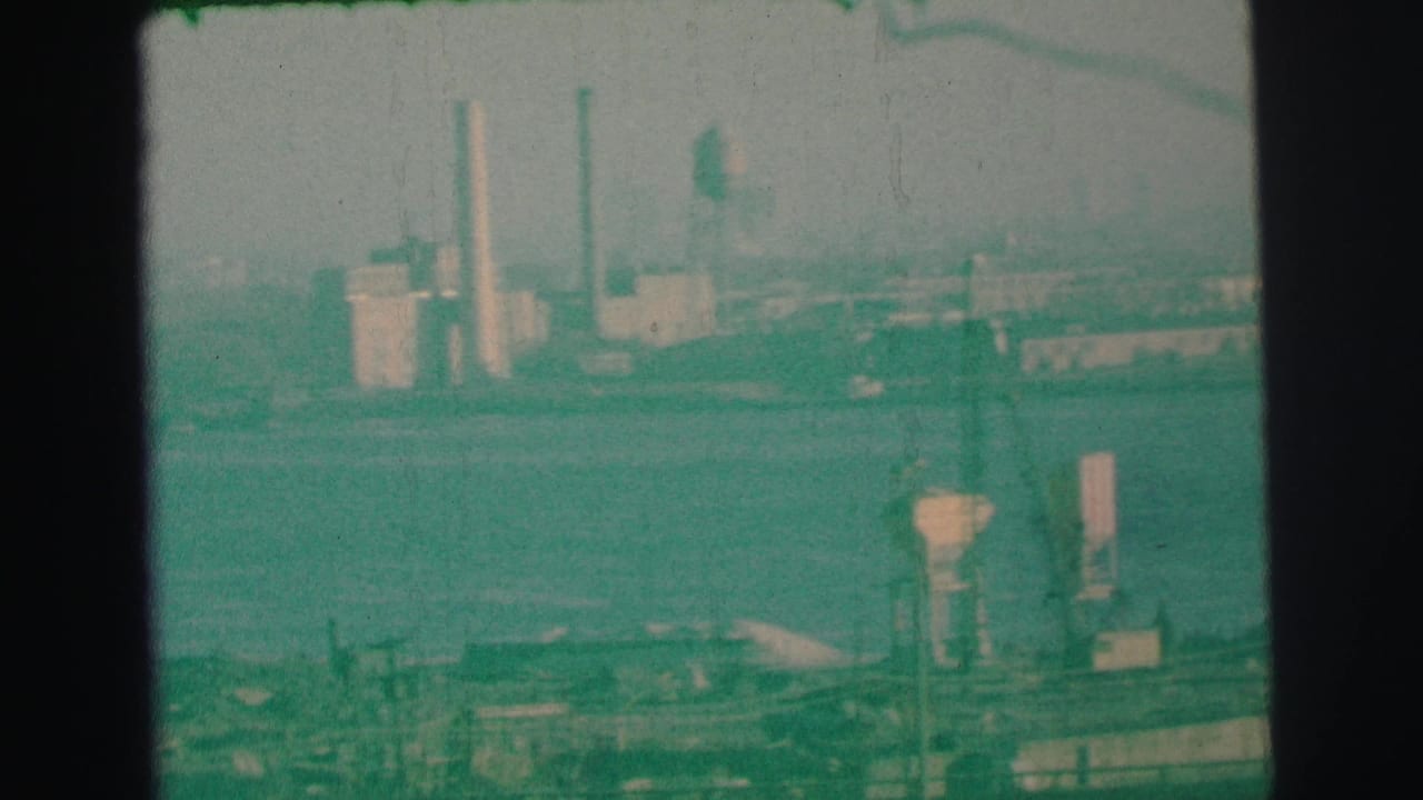 1973 Lynbrook, New York 8mm frame showing a hazy industrial shoreline with smokestacks, low buildings, and foreground structures across water.