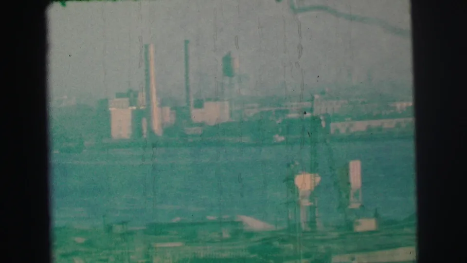 1973 Lynbrook, New York 8mm frame showing a hazy factory shoreline, smokestacks, water, and dark gate borders from the scan.