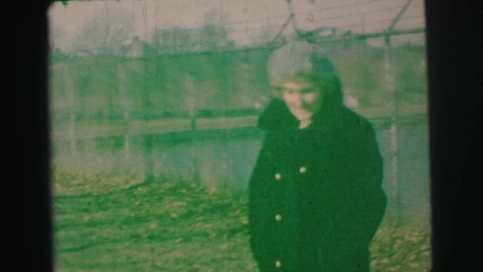 In Lynbrook, New York, a woman in a dark coat and light hat stands near lake water, fencing, and bare trees in 1973 home-movie footage.