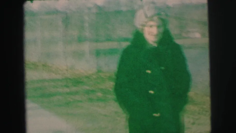 A woman in a dark coat and light hat faces the camera beside the lake in Lynbrook, with soft focus and green-shifted 8mm color.