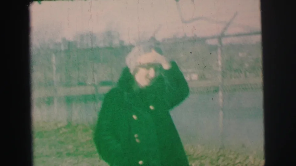 The woman shades her eyes with one hand while standing near the lake and fence in 1973 Lynbrook home-movie footage.
