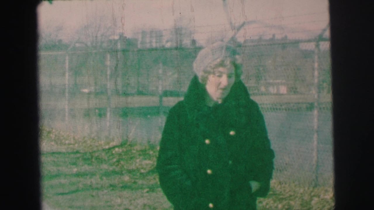 Woman in a dark winter coat and light hat stands in a fenced yard.