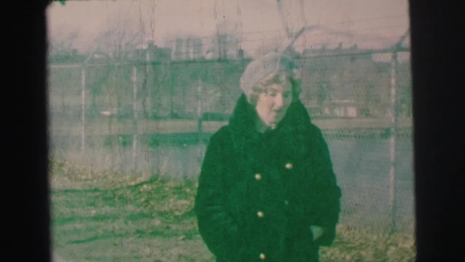 In 1973 Lynbrook home-movie footage, a woman in a dark green winter coat and light hat faces the camera outdoors by a chain-link fence.