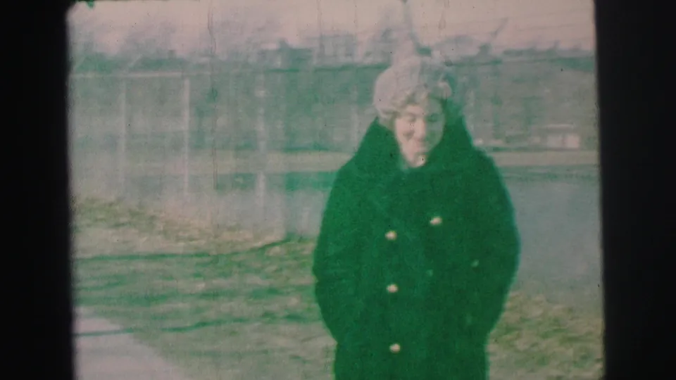 The woman remains facing the camera in a Lynbrook yard, wearing a heavy dark coat and pale hat against a soft, faded 8mm background.