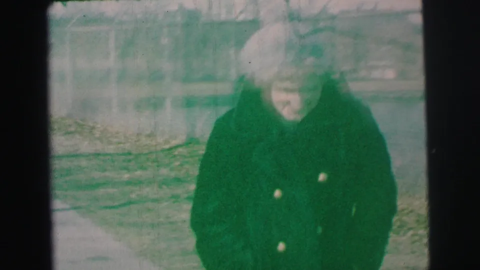 A closer, blurred frame shows the woman in her winter coat looking downward as she moves across the yard in the 1973 home movie.