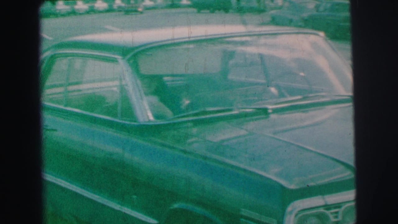 The parked sedan remains in frame near the curb, showing chrome trim, windshield, and soft 1970s 8mm home-movie texture.