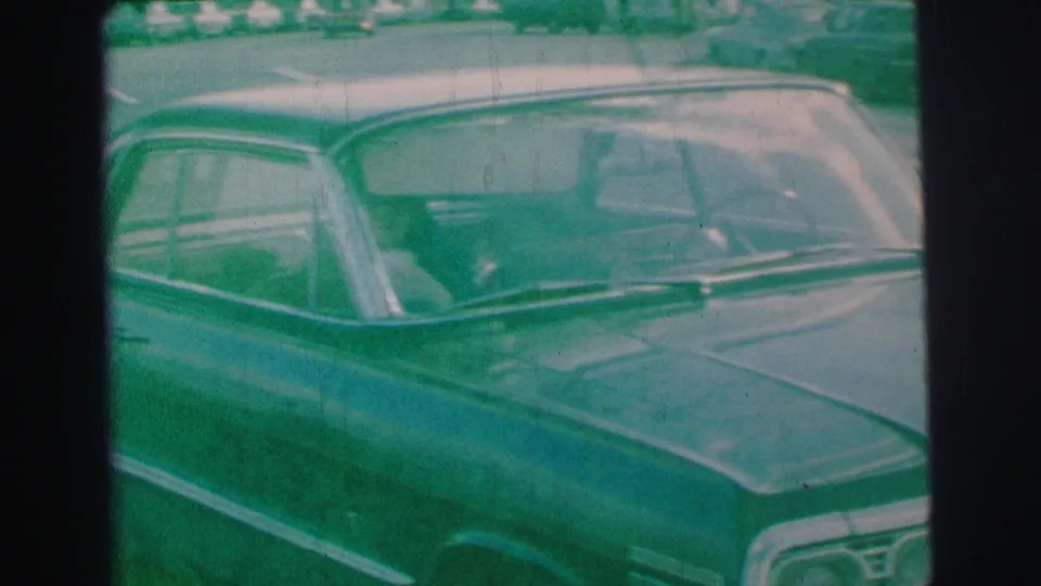 The parked sedan remains in frame near the curb, showing chrome trim, windshield, and soft 1970s 8mm home-movie texture.