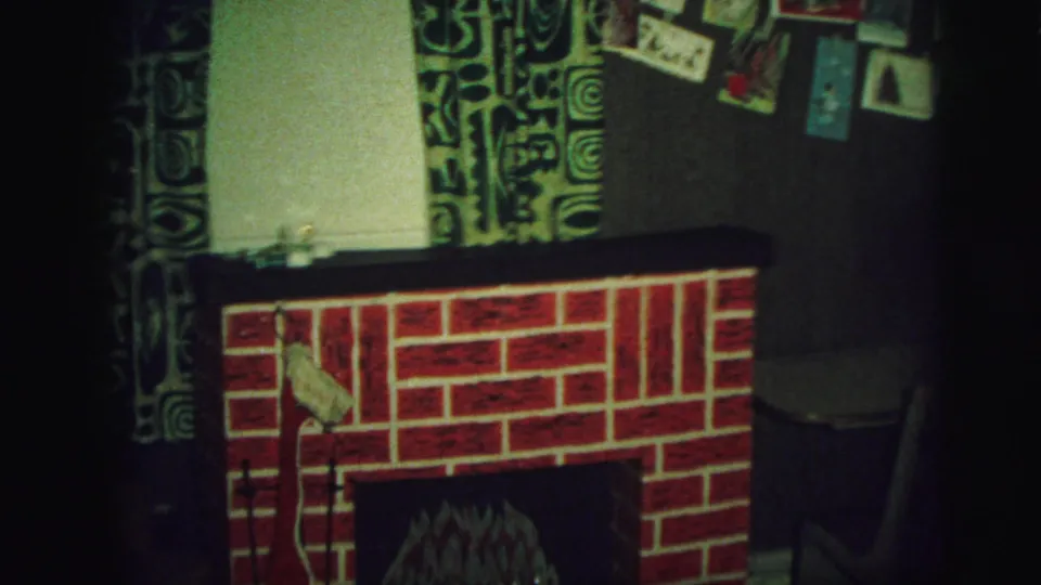 1973 Lynbrook home-movie frame showing a red fake fireplace, patterned curtains, pinned postcards, and dim living-room lighting.