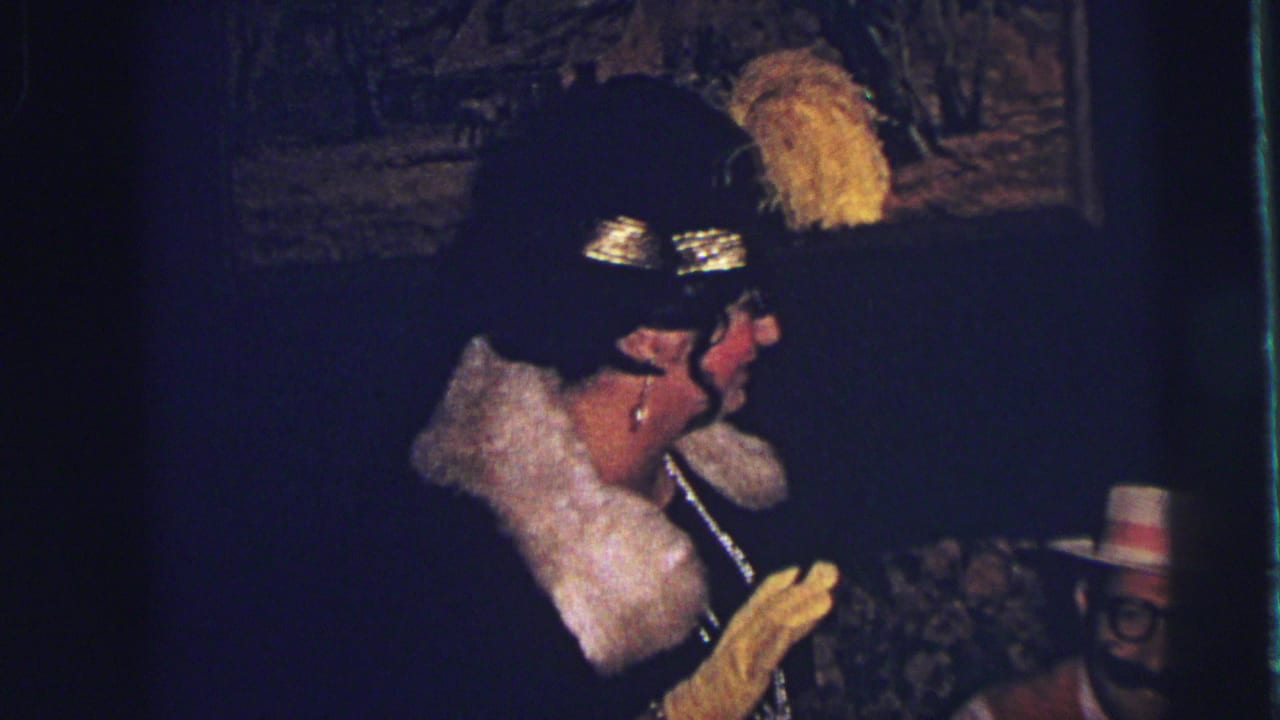In a 1973 Lynbrook home-movie frame, a woman in a feathered black hat, pale collar, gloves, and necklace stands near framed wall art.