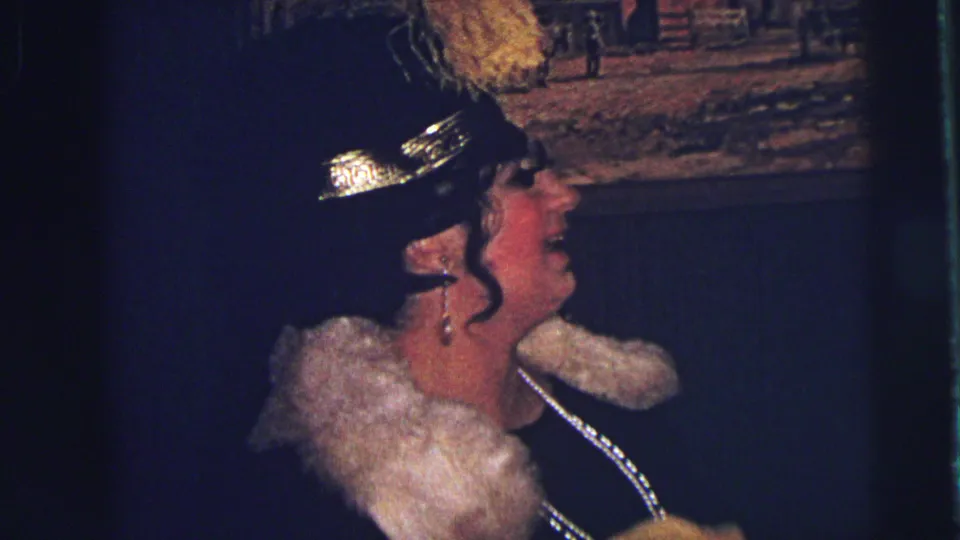The woman in a black feathered hat and pale fur-like collar smiles in profile in warm, grainy 1973 Lynbrook home-movie footage.