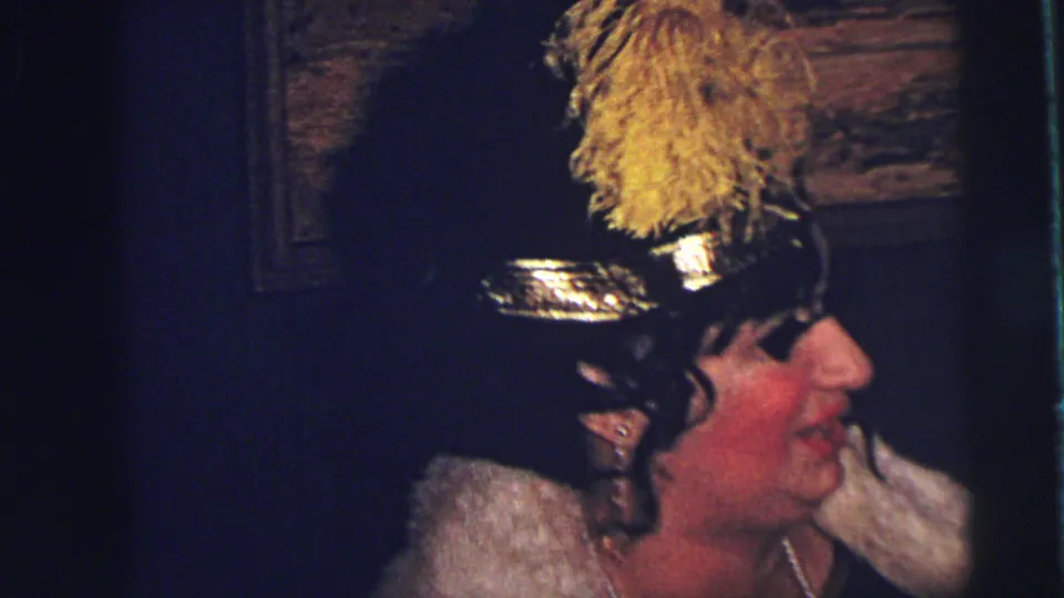 A closer 1973 Lynbrook frame shows the woman in a feathered black hat, pale collar, yellow gloves, and necklace under dim indoor light.
