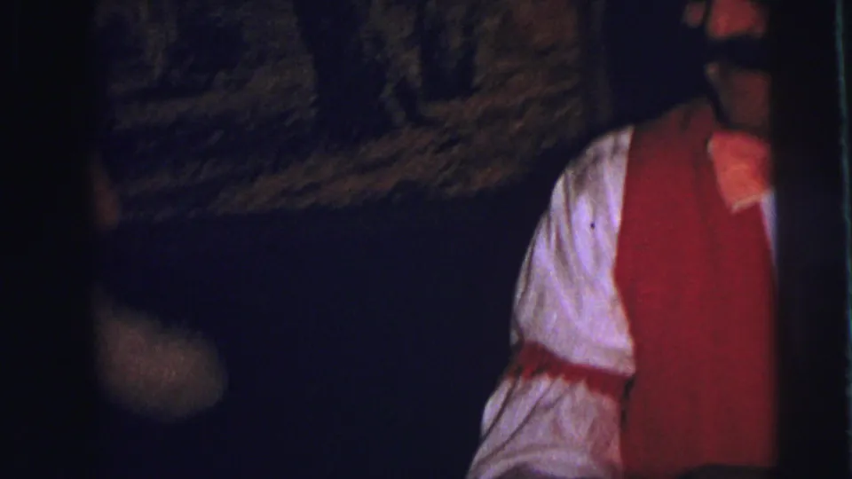 A man in a red vest, white shirt, bow tie, glasses, and fake mustache appears in the background of the 1973 Lynbrook party footage.