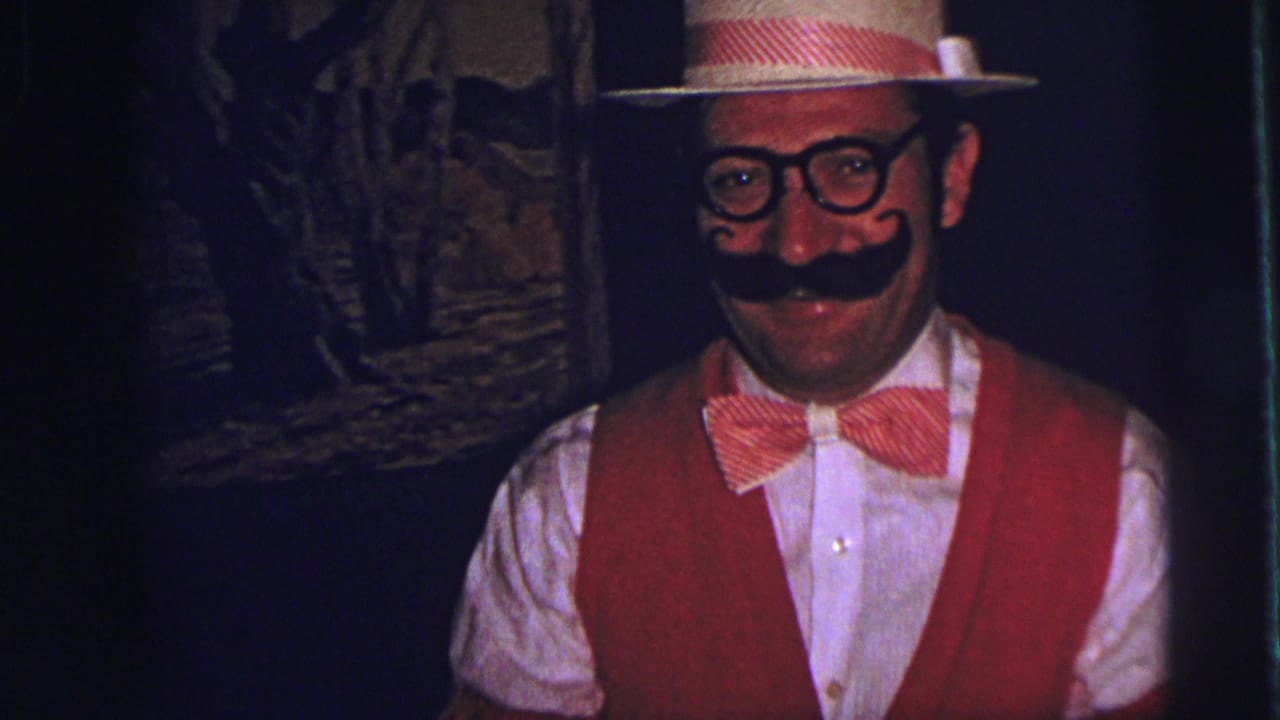 Costumed man in red vest, bow tie, straw hat, fake mustache, and glasses smiling at the camera.