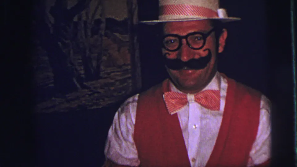 In 1973 Lynbrook home-movie footage, a costumed man in a red vest and straw hat smiles toward the camera in a dim interior.