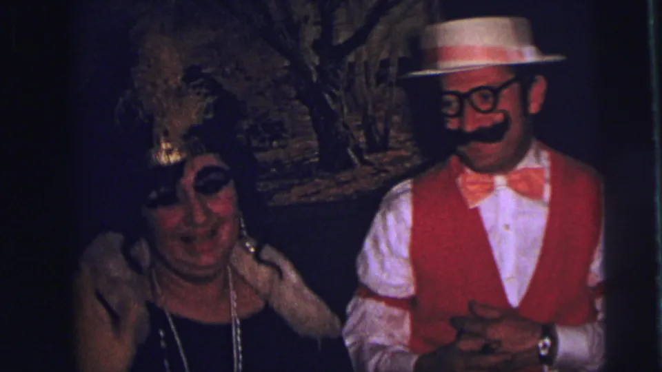 A woman in a feathered headpiece and pearls sits near a costumed man in red vest and straw hat in 1973 Lynbrook footage.