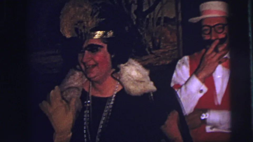 In a dim 1973 Lynbrook interior, a woman in a feathered headpiece smiles while a man in carnival costume stands behind her.