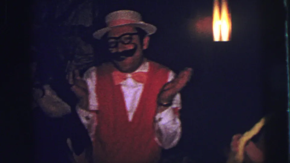 A costumed man in red vest, bow tie, straw hat, fake mustache, and glasses gestures playfully in 1973 Lynbrook footage.