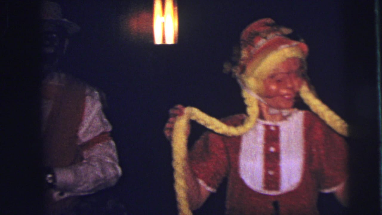 Costumed woman holding long yellow braids while a man in a red vest stands nearby in dim light.