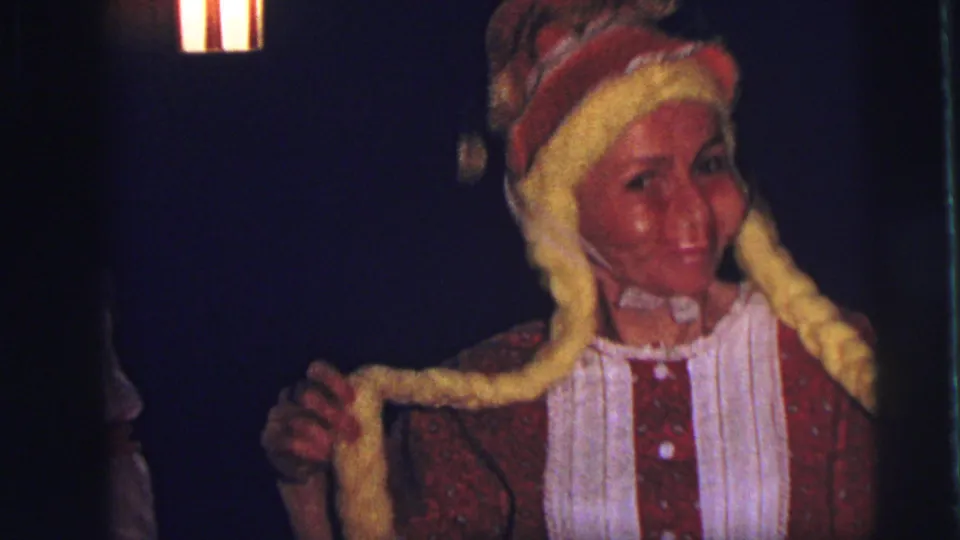 A smiling woman in a red German-style costume and yellow braided hat appears in close view in 1973 Lynbrook home-movie footage.