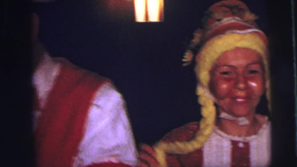 A costumed woman smiles beside a cropped performer in a red vest during a dimly lit 1973 Lynbrook home-movie dance scene.